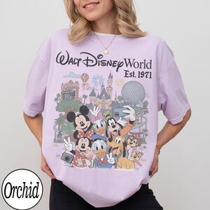 May include: A light purple short-sleeve t-shirt featuring a graphic print of Walt Disney World Est. 1971 with Mickey Mouse, Minnie Mouse, Donald Duck, Daisy Duck, Goofy, Pluto, Chip, and Dale in front of park landmarks and fireworks.