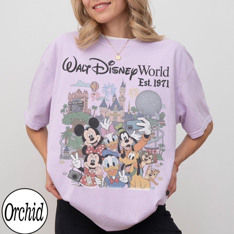 May include: A light purple short-sleeve t-shirt featuring a graphic print of Walt Disney World Est. 1971 with Mickey Mouse, Minnie Mouse, Donald Duck, Daisy Duck, Goofy, Pluto, Chip, and Dale in front of park landmarks and fireworks.