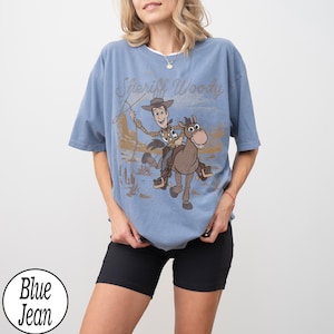 May include: A blue t-shirt featuring a graphic of Sheriff Woody riding a horse with the text "Sheriff Woody" in a distressed font. The shirt has short sleeves and a relaxed fit. A white circle with the words "Blue Jean" is in the bottom left corner.