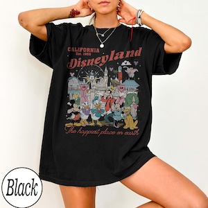 May include: Black t-shirt with a Disneyland graphic. The design features the text "CALIFORNIA Est. 1955 Disneyland" above a colourful illustration of Disney characters and landmarks. The phrase "The happiest place on earth" is below the illustration.