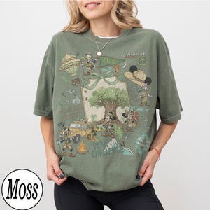May include: Olive green t-shirt with a safari-themed cartoon graphic, including a hot air balloon and jeep. The design features the text "Kilimanjaro" and "Adventure Awaits". The shirt has a loose fit and is made of soft material.