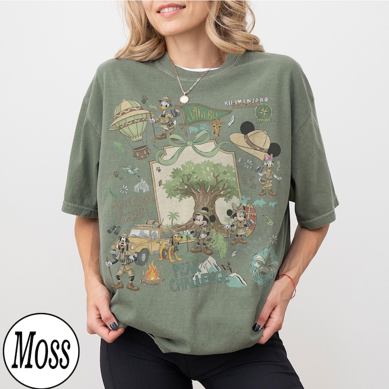 May include: Olive green t-shirt with a safari-themed cartoon graphic, including a hot air balloon and jeep. The design features the text "Kilimanjaro" and "Adventure Awaits". The shirt has a loose fit and is made of soft material.