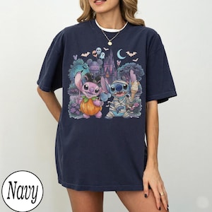 May include: A navy blue short-sleeve t-shirt featuring a graphic print of two blue and pink alien characters. One character holds a pumpkin, while the other is wrapped like a mummy and holds an ice cream cone. A castle and bats are in the background.