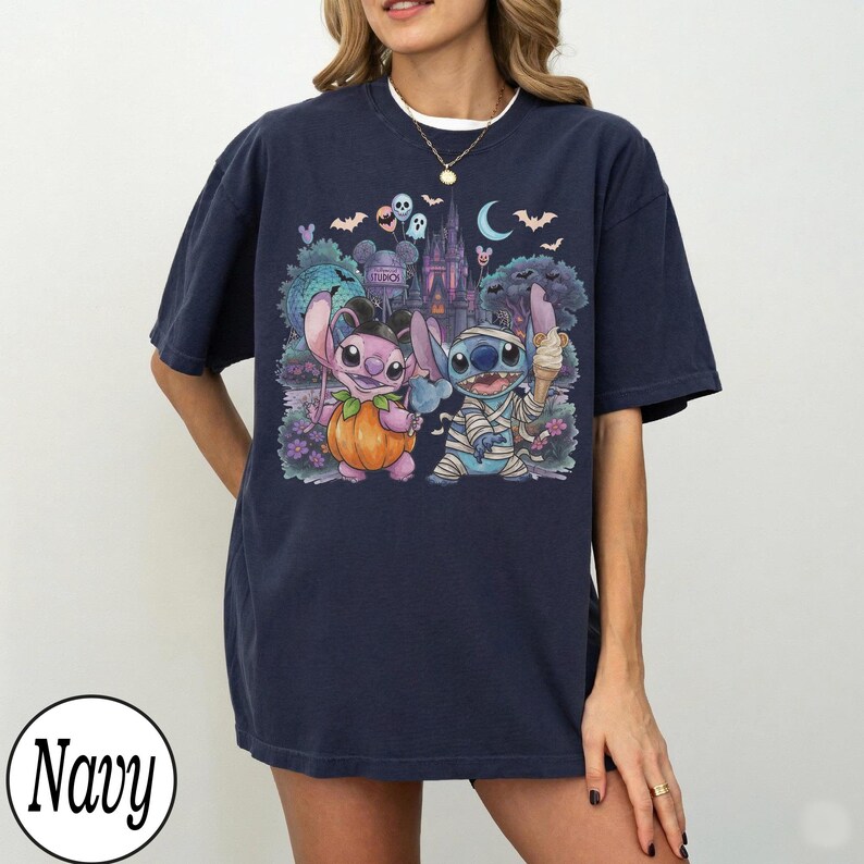 May include: A navy blue short-sleeve t-shirt featuring a graphic print of two blue and pink alien characters. One character holds a pumpkin, while the other is wrapped like a mummy and holds an ice cream cone. A castle and bats are in the background.