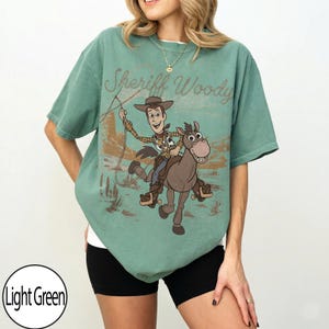 May include: Light green t-shirt featuring a graphic of Sheriff Woody riding a horse, with the text "Sheriff Woody" above the graphic. The shirt is a relaxed fit and has short sleeves.