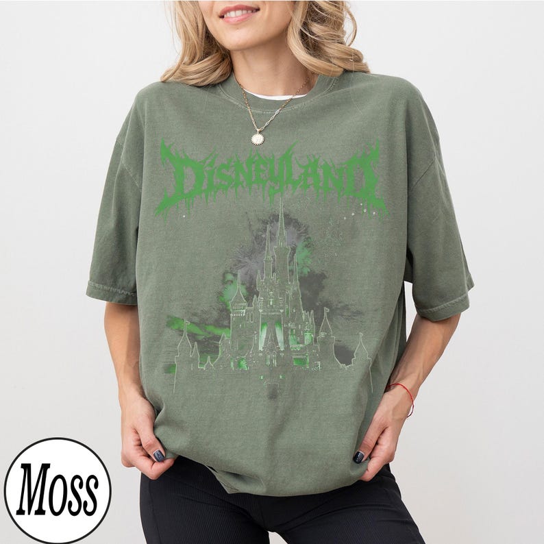 May include: Olive green t-shirt with the word "DISNEYLAND" in green, stylized text. Below the text is a graphic of a castle in shades of gray and green. The shirt has short sleeves and a relaxed fit.