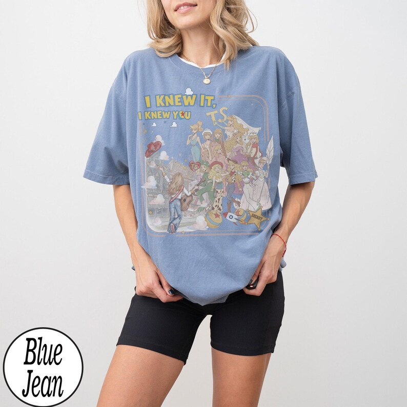 May include: A dusty blue short-sleeve t-shirt featuring a graphic print of various animated characters and the text "I KNEW IT, I KNEW YOU" along with "T.S." in a rectangular frame. The shirt has a crew neckline and a loose, relaxed fit.