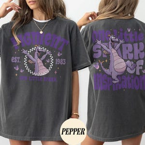 May include: Dark gray t-shirt featuring a purple dragon graphic with the text "Figment" and "One Little Spark of Inspiration." The front design includes "est. 1983." The shirt has a relaxed fit and short sleeves.