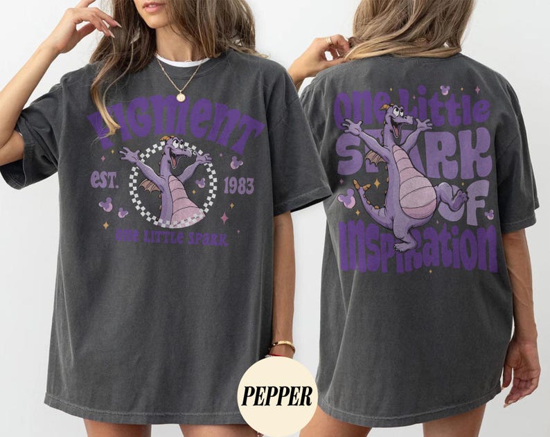 May include: Dark gray t-shirt featuring a purple dragon graphic with the text "Figment" and "One Little Spark of Inspiration." The front design includes "est. 1983." The shirt has a relaxed fit and short sleeves.