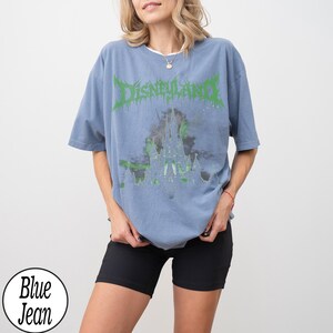 May include: A blue t-shirt with the word "Disneyland" in green, stylized text above a castle graphic. The shirt is a light blue color and has short sleeves. The model is wearing black shorts.