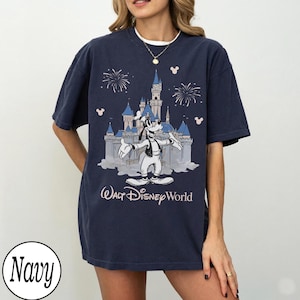 May include: Navy blue t-shirt with a graphic of Goofy in front of Cinderella's castle, fireworks, and Mickey Mouse icons. The text "Walt Disney World" is below the castle. The shirt has a relaxed fit.