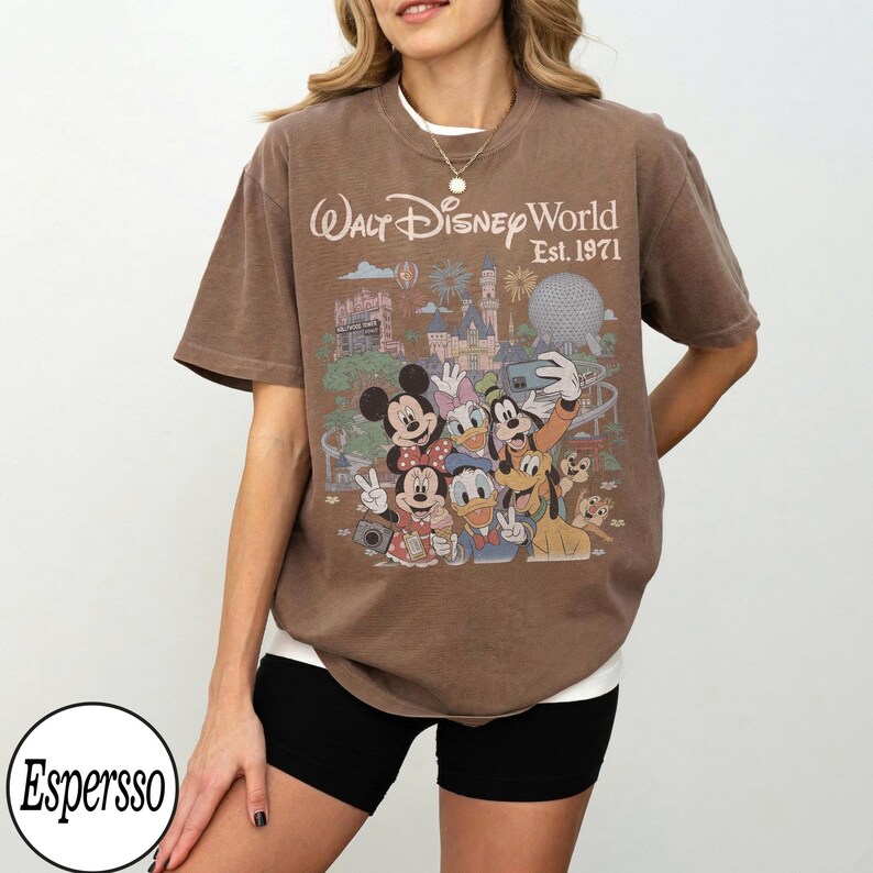 May include: A brown short-sleeve t-shirt featuring a graphic print of Disney characters including Mickey Mouse, Minnie Mouse, Donald Duck, Daisy Duck, Goofy, and Pluto. The text reads Walt Disney World Est. 1971 with park landmarks.