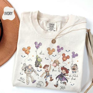 Pode incluir: Camiseta de cor marfim com um gr&aacute;fico dos personagens de Toy Story da Disney segurando bal&otilde;es em forma de Mickey Mouse. O design inclui Buzz Lightyear, Woody, Jessie e Sr. Cabe&ccedil;a de Batata. A camisa tem a etiqueta da marca Comfort Colors.