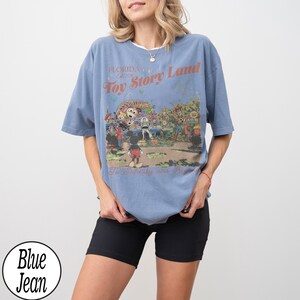 May include: A blue t-shirt featuring a graphic of Toy Story Land in Florida. The design includes Mickey Mouse and other characters. The text on the shirt reads "Florida Toy Story Land" and "To Infinity and Beyond."
