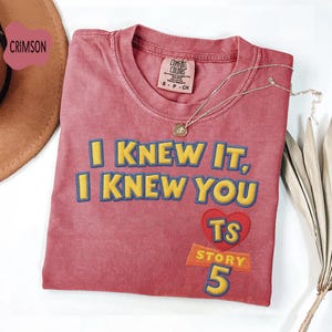 Puede incluir: Una camiseta de manga corta color carmes&iacute; con el texto bordado en amarillo I KNEW IT, I KNEW YOU, un coraz&oacute;n rojo con las letras amarillas TS y el texto amarillo STORY 5. La camiseta tiene una etiqueta Comfort Colors en el cuello.