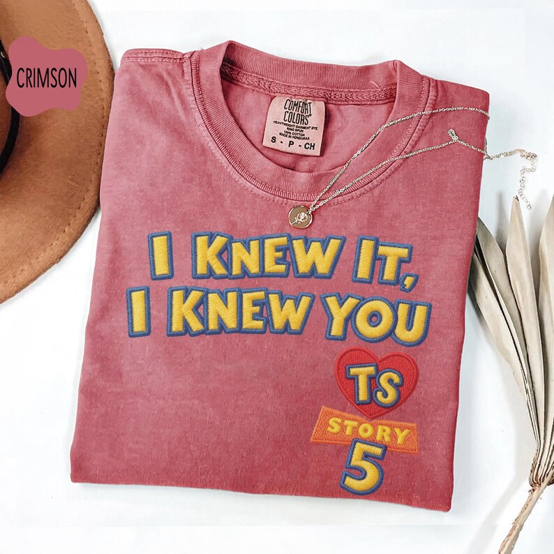 Puede incluir: Una camiseta de manga corta color carmes&iacute; con el texto bordado en amarillo I KNEW IT, I KNEW YOU, un coraz&oacute;n rojo con las letras amarillas TS y el texto amarillo STORY 5. La camiseta tiene una etiqueta Comfort Colors en el cuello.