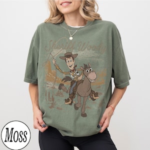 May include: Green t-shirt featuring a graphic of Sheriff Woody riding a horse with the text "Sheriff Woody". The design is in a vintage style. The shirt has short sleeves and a crew neck.