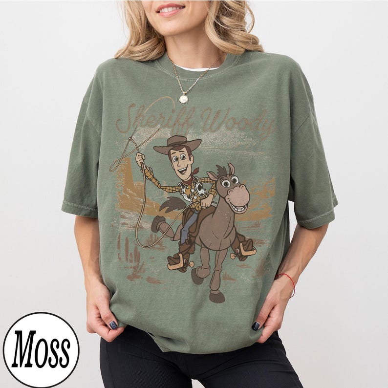 May include: Green t-shirt featuring a graphic of Sheriff Woody riding a horse with the text "Sheriff Woody". The design is in a vintage style. The shirt has short sleeves and a crew neck.