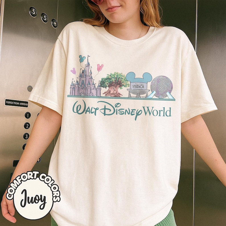 Puede incluir: Camiseta color crema con un gr&aacute;fico verde azulado de los lugares emblem&aacute;ticos de Disney World, incluyendo un castillo, un &aacute;rbol y Spaceship Earth. El texto "Walt Disney World" est&aacute; impreso debajo. La camiseta tambi&eacute;n tiene el texto "COMFORT COLORS Juoy" en la parte inferior izquierda.
