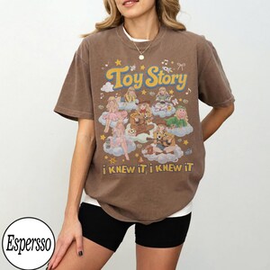 Może przedstawiać: Brązowy t-shirt z kr&oacute;tkim rękawem z nadrukiem graficznym zawierającym tekst Toy Story oraz i KNEW iT, i KNEW iT. Wz&oacute;r przedstawia wiele ilustrowanych postaci siedzących na chmurach, otoczonych małymi gwiazdkami, nutami i kokardkami.