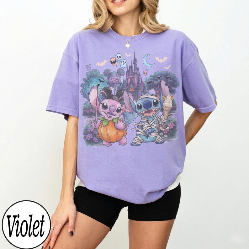 May include: A light violet short-sleeved t-shirt featuring a graphic print of two blue and pink alien characters. One character is dressed as a pumpkin and the other as a mummy, set against a background of a castle, trees, and bats.