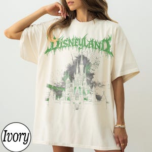 May include: Ivory-colored t-shirt with a green and gray graphic of a castle and the word "Disneyland" in green, stylized text. The shirt has short sleeves and a relaxed fit, suitable for casual wear.