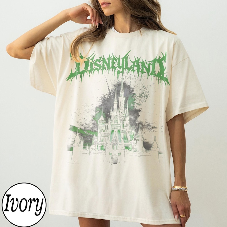 May include: Ivory-colored t-shirt with a green and gray graphic of a castle and the word "Disneyland" in green, stylized text. The shirt has short sleeves and a relaxed fit, suitable for casual wear.