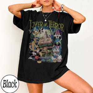 K&ouml;nnte beinhalten: Schwarzes T-Shirt mit einem Aufdruck des Tower of Terror, Mickey Mouse, Minnie Mouse, Donald Duck und Goofy. Der Text "Tower of Terror" ist gr&uuml;n, mit "Hollywood Tower Hotel" darunter.