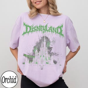May include: A light purple t-shirt with the word "Disneyland" in green, stylized text. Below the text is a graphic of a castle in green and gray. The shirt has short sleeves and a crew neck. The brand name "Orchid" is printed on the bottom left.