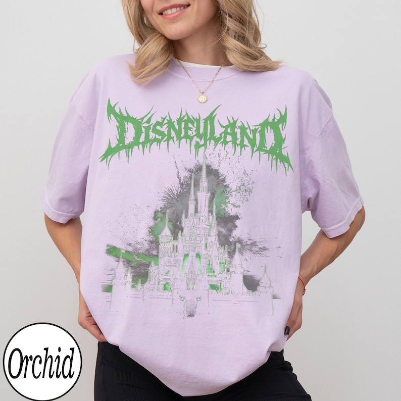 May include: A light purple t-shirt with the word "Disneyland" in green, stylized text. Below the text is a graphic of a castle in green and gray. The shirt has short sleeves and a crew neck. The brand name "Orchid" is printed on the bottom left.