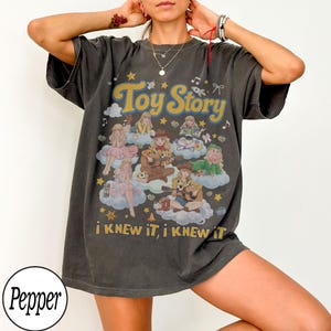 Może przedstawiać: Ciemnoszary t-shirt z kr&oacute;tkim rękawem z nadrukiem postaci z Toy Story siedzących na chmurach. Wz&oacute;r zawiera napis Toy Story oraz i KNEW iT, i KNEW iT ż&oacute;łtymi literami, otoczony gwiazdkami, nutami muzycznymi i małymi kokardkami.