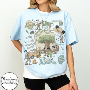 May include: Pale blue t-shirt with a vibrant graphic of cartoon characters on safari. The design includes a hot air balloon, a jeep, and the text "Adventure Awaits!" and "Peak Challenge."