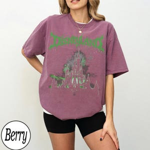 May include: Berry-colored t-shirt with the word "DISNEYLAND" in green, stylized text. The shirt features a graphic of a castle in green and gray. The model is wearing black shorts and a white undershirt.