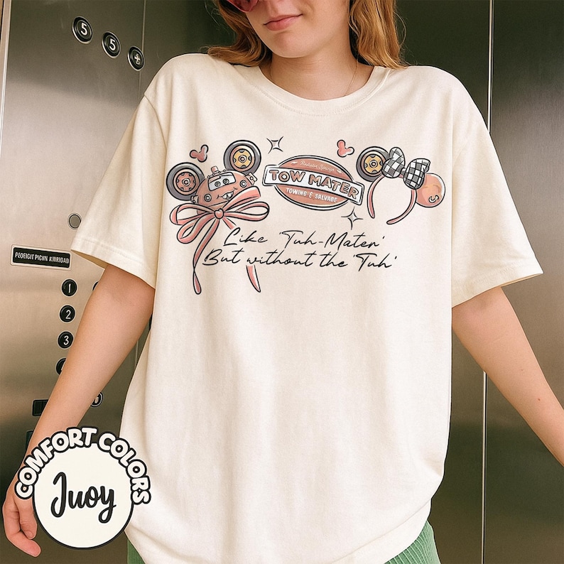 Puede incluir: Camiseta color crema con un dise&ntilde;o de Tow Mater, un lazo rosa y el texto "Like Tuh-Mater But without the Tuh." La camiseta tambi&eacute;n tiene un logotipo de Comfort Colors.