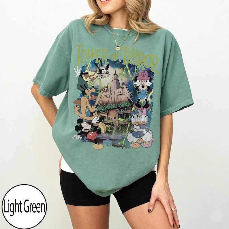 K&ouml;nnte beinhalten: Hellgr&uuml;nes T-Shirt mit einem Aufdruck des Hollywood Tower Hotels, Mickey Mouse, Minnie Mouse, Donald Duck und Goofy. Der Text "Tower of Terror" befindet sich &uuml;ber dem Aufdruck. Das Shirt hat eine lockere Passform.