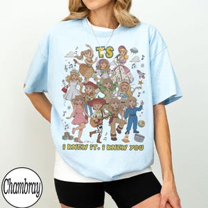 May include: A light blue t-shirt featuring a graphic print of multiple cartoon characters in various outfits, including a space suit and a cowgirl costume. The text TS is at the top, and the text I KNEW IT, I KNEW YOU is at the bottom.