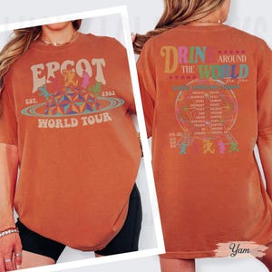 May include: Burnt orange t-shirt with "EPCOT World Tour" graphic on the front, featuring a colorful geometric design and the year 1982. The back of the shirt says "DRIN AROUND THE WORLD The Tour" with a globe graphic and country names.