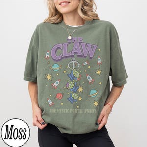 Puede incluir: Una camiseta de manga corta color verde musgo con un estampado gr&aacute;fico de tres alien&iacute;genas verdes colgando de una garra mec&aacute;nica. El texto dice THE CLAW y THE MYSTIC PORTAL AWAITS, rodeado de ilustraciones de cohetes, planetas y estrellas.