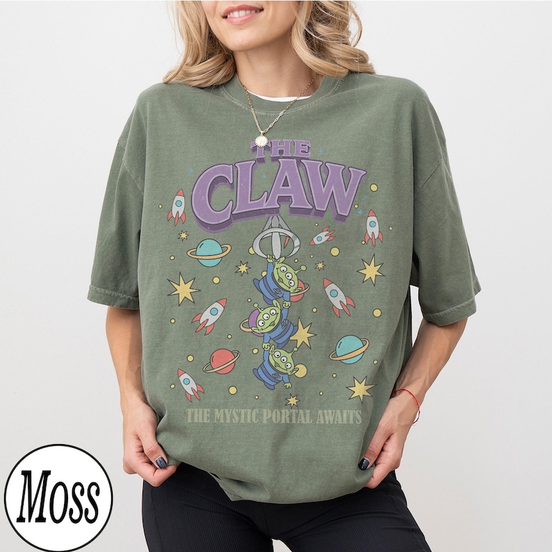 Puede incluir: Una camiseta de manga corta color verde musgo con un estampado gr&aacute;fico de tres alien&iacute;genas verdes colgando de una garra mec&aacute;nica. El texto dice THE CLAW y THE MYSTIC PORTAL AWAITS, rodeado de ilustraciones de cohetes, planetas y estrellas.