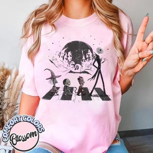 Peut inclure: T-shirt rose clair avec un motif de personnages de L'&Eacute;trange No&euml;l de Monsieur Jack traversant une rue, avec une pleine lune et une ville effrayante en arri&egrave;re-plan. Le t-shirt porte les mots "Comfort Colors Blossom" dans un cercle en bas.