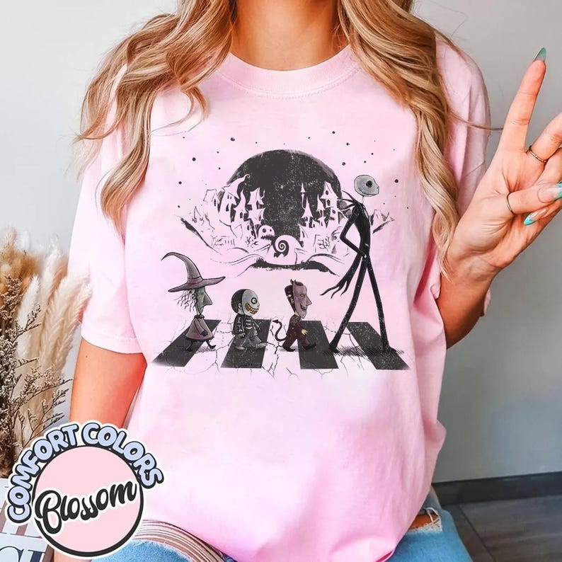 Peut inclure: T-shirt rose clair avec un motif de personnages de L'&Eacute;trange No&euml;l de Monsieur Jack traversant une rue, avec une pleine lune et une ville effrayante en arri&egrave;re-plan. Le t-shirt porte les mots "Comfort Colors Blossom" dans un cercle en bas.