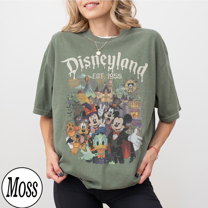 Puede incluir: Una camiseta de manga corta color verde musgo con un estampado gr&aacute;fico de personajes de Disney con disfraces de Halloween. El texto dice Disneyland EST. 1955. El dise&ntilde;o incluye a Mickey, Minnie, Goofy, el Pato Donald y Pluto frente a un castillo.