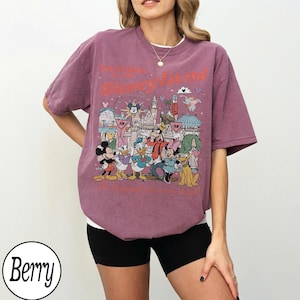 May include: Berry-coloured t-shirt featuring a Disneyland graphic with Mickey Mouse, Donald Duck, and other Disney characters. The graphic includes the Disneyland castle and other park icons. The shirt has a crew neck and short sleeves.