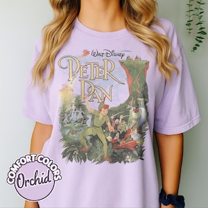 May include: A light purple Comfort Colors t-shirt featuring a graphic from the Disney movie Peter Pan. The graphic includes the movie title and characters like Peter Pan, Captain Hook, and Tinkerbell. The shirt is a solid color with a round logo that says "Comfort Colors Orchid".
