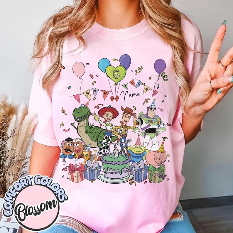 May include: Pink t-shirt featuring a colorful graphic of Toy Story characters celebrating a birthday. The design includes a cake, balloons, and the number 5. The shirt also has the text "Name" and "Comfort Colors Blossom".