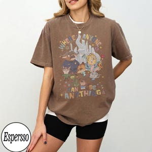 May include: Brown t-shirt with a colourful graphic featuring cartoon characters and the text "WHERE ANYONE CAN BE ANYTHING." The shirt has a relaxed fit and short sleeves. The design includes various animal characters in a whimsical style.