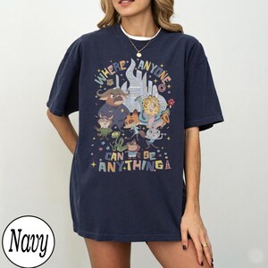 May include: Navy blue t-shirt with a colourful graphic that reads "WHERE ANYONE CAN BE ANYTHING." The design features cartoon characters and a city skyline. The t-shirt has a relaxed fit.