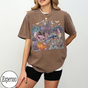 May include: A brown short-sleeve t-shirt featuring a graphic print of two blue and pink alien characters dressed in Halloween costumes. One character is inside a pumpkin, and the other is wrapped in bandages like a mummy, with a castle in the background.