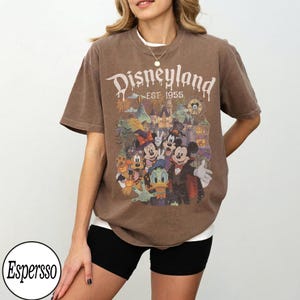 Puede incluir: Una camiseta marr&oacute;n de manga corta con el texto Disneyland EST. 1955 en color blanco. El gr&aacute;fico incluye ilustraciones coloridas de Mickey Mouse, Minnie Mouse, Donald Duck y Pluto vestidos con disfraces de Halloween cerca de un castillo.