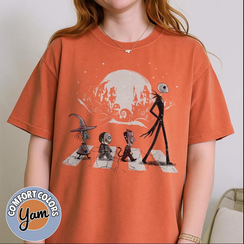Peut inclure: T-shirt orange avec un motif de personnages de L'&Eacute;trange No&euml;l de Monsieur Jack traversant un passage pi&eacute;ton devant une pleine lune. Le t-shirt porte les logos Comfort Colors et Yam.
